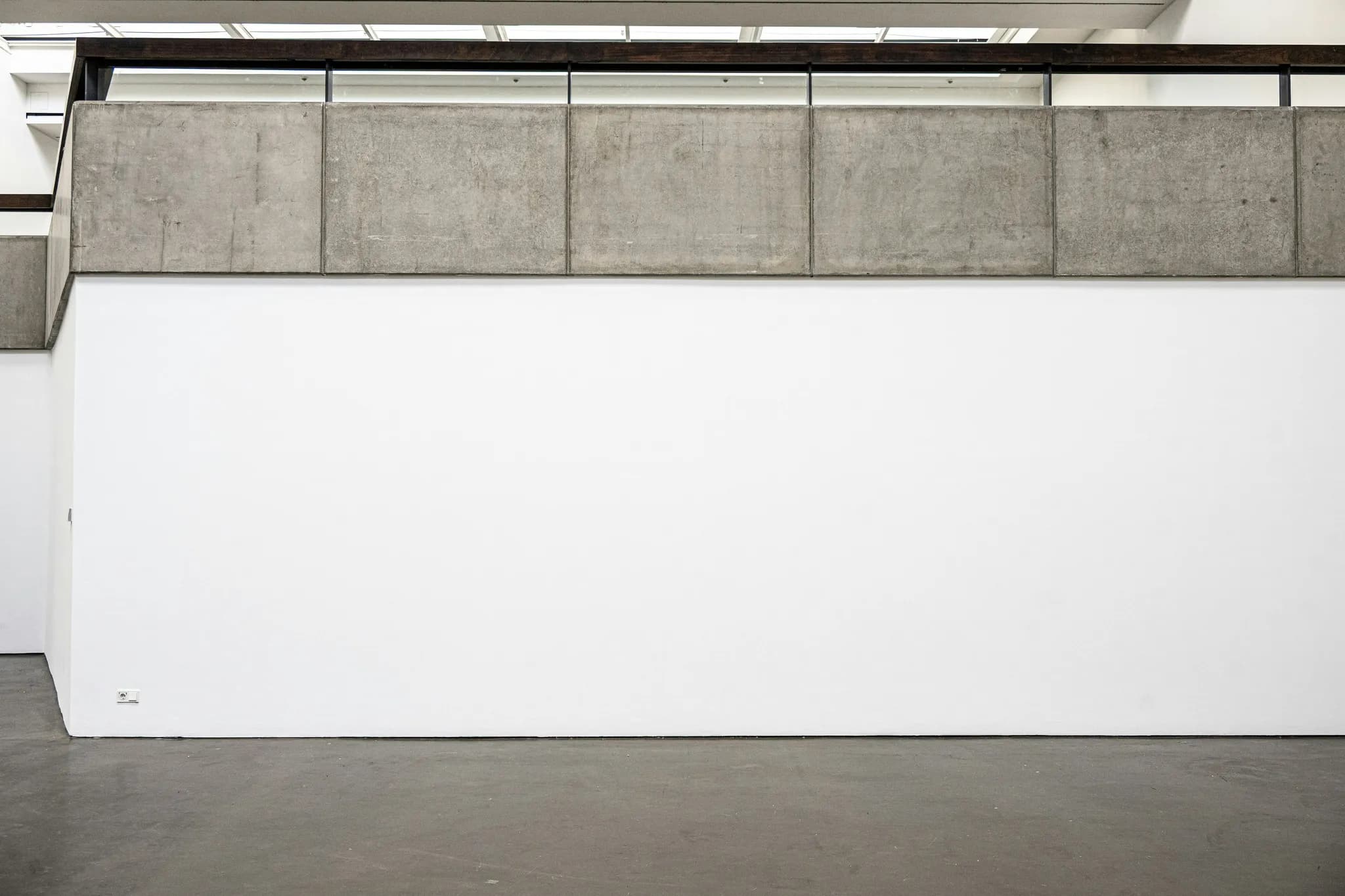 A tall white gallery wall under a concrete parapet, above a grey concrete floor