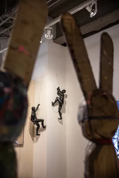 Climbing figures by Ancizar Marin on a gallery wall, seen past a wooden sculpture in the foreground