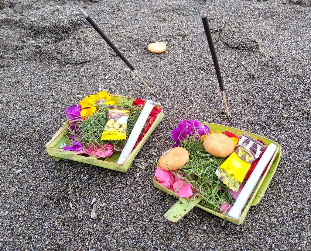 Two canang sari offerings with flowers and incense on a black sand beach in Bali
